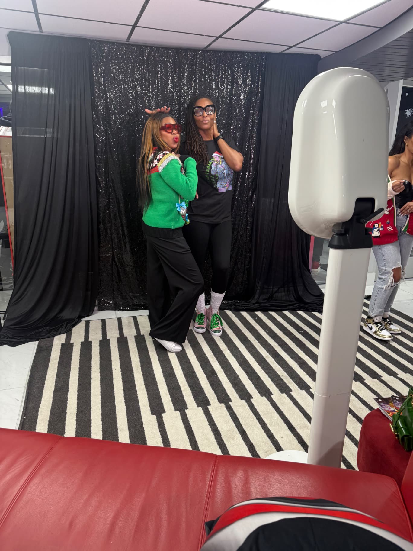 Two guests in holiday sweaters posing at a black sequin backdrop booth